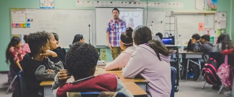 Students at their desks listen to a teacher at the front of the classroom