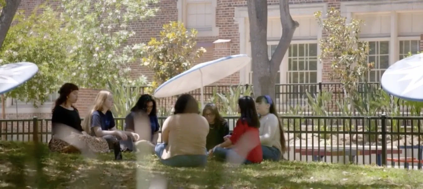 A group of high school students sits in a circle on the grass outside their school during a mindfulness practice
