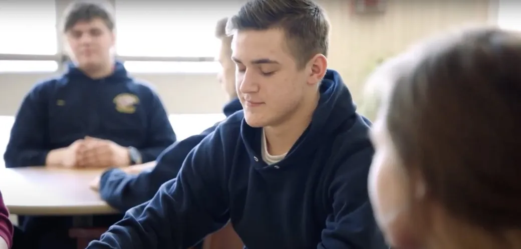 A high school student sits at a classroom table with eyes closed during a mindfulness practice.