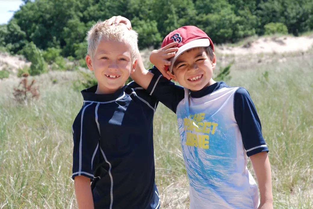 Two smiling boys stand arm in arm in a sunny grassy field
