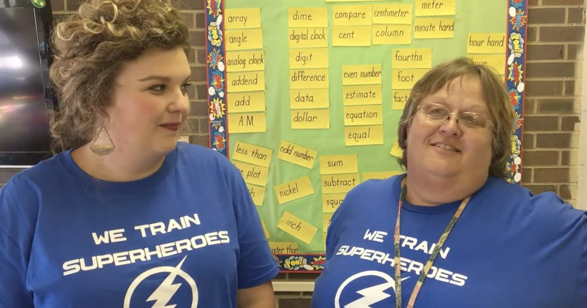 Two educators in matching blue shirts stand in front of a math vocabulary board
