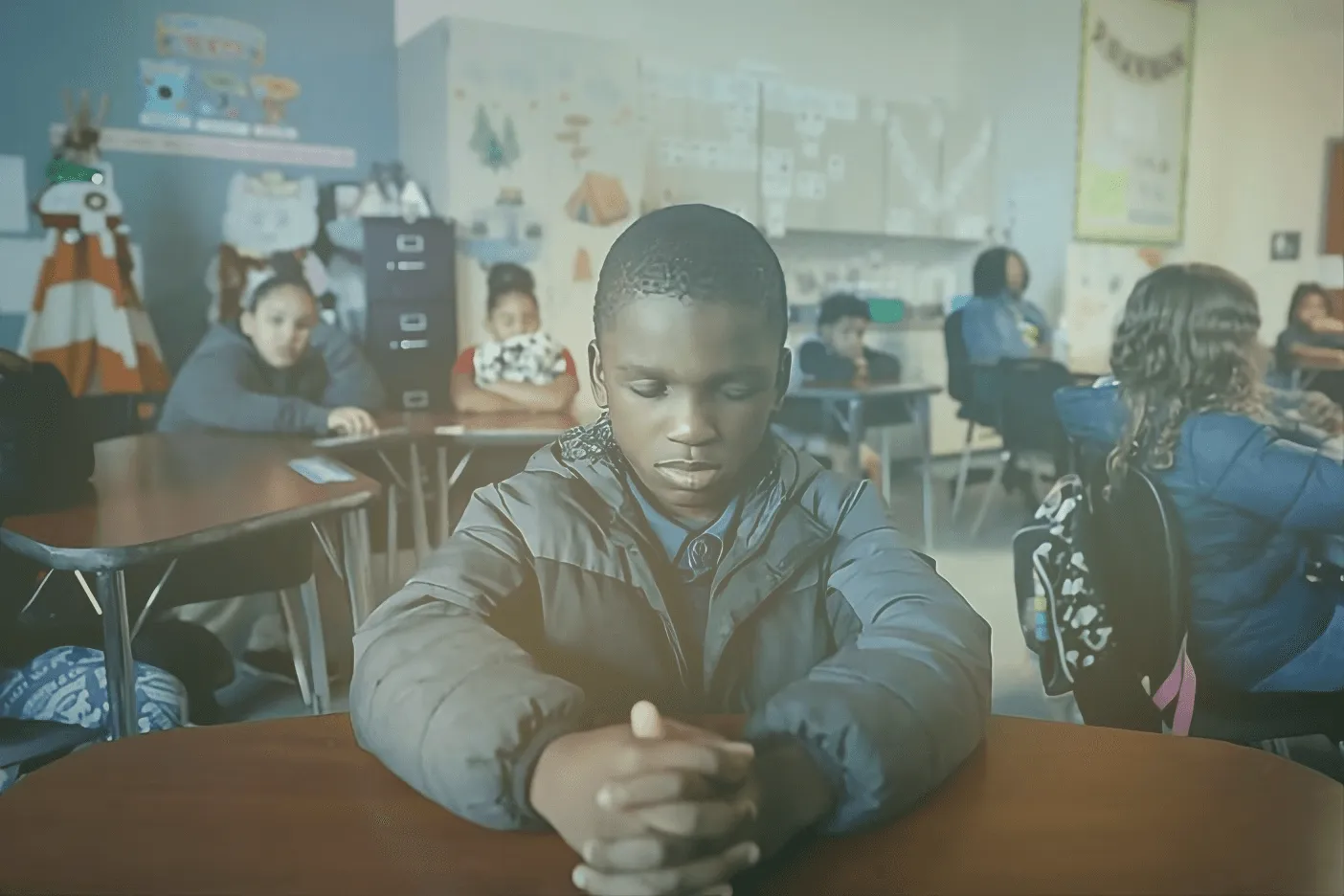 A student sits quietly with lowered eyes during a calm moment in a busy classroom.