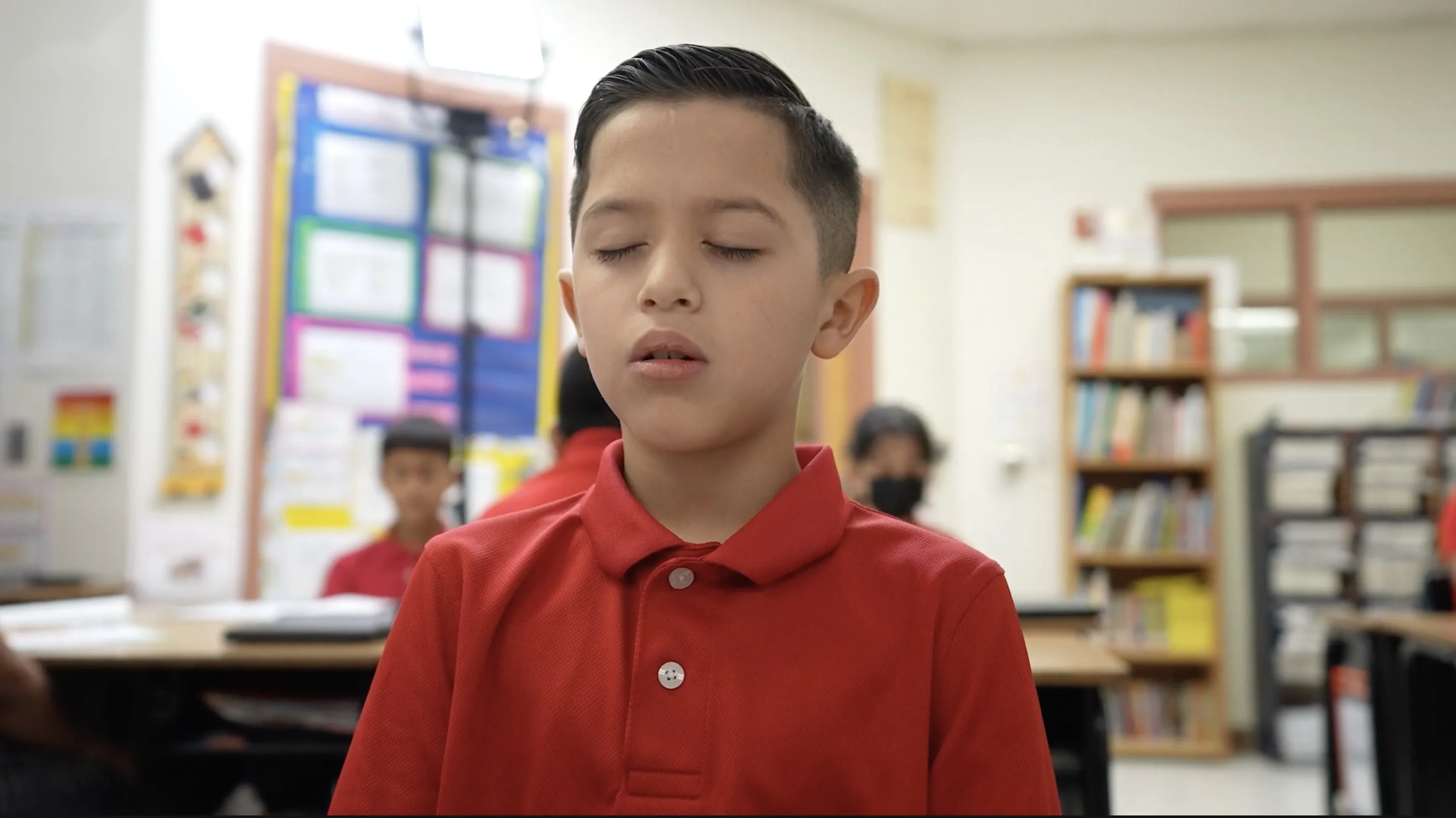 A student in a red school polo sits with eyes closed during a daily mindfulness practice in his classroom