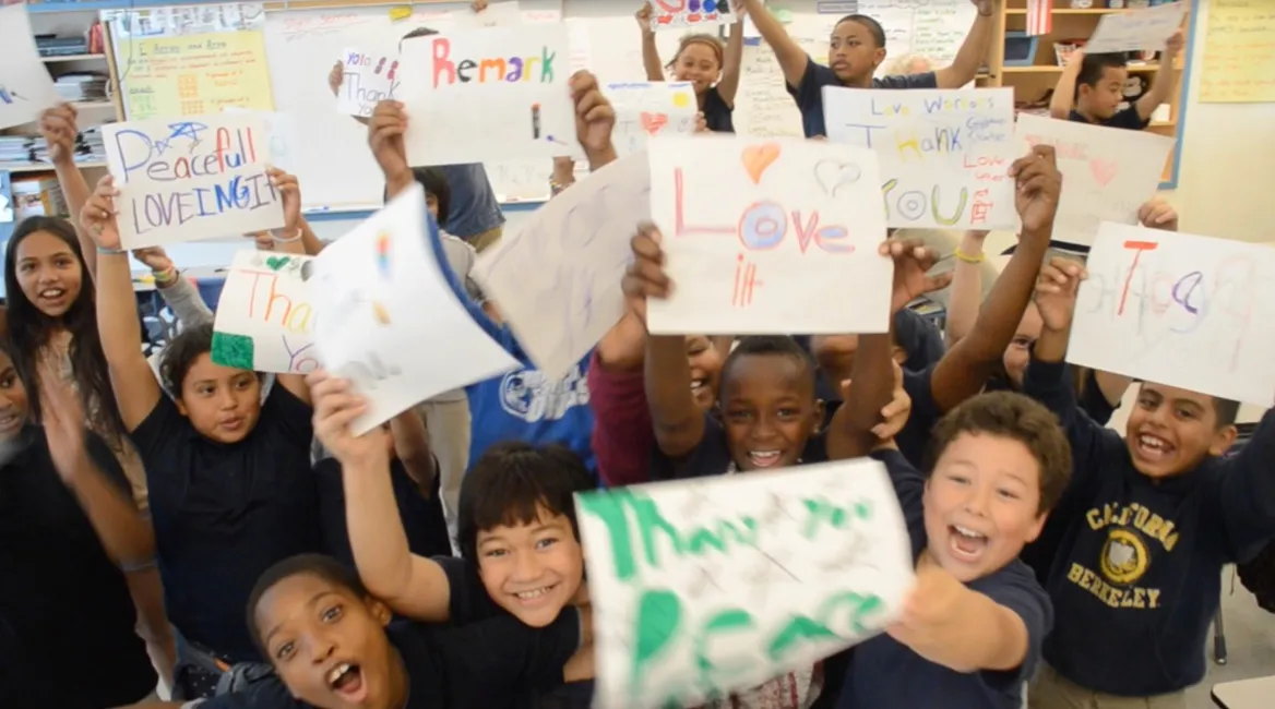Smiling students hold up handmade thank-you and peace signs in their classroom