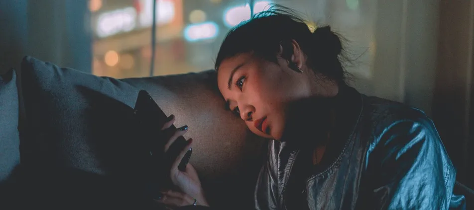 A young woman rests her head on her hand, lit by her phone screen