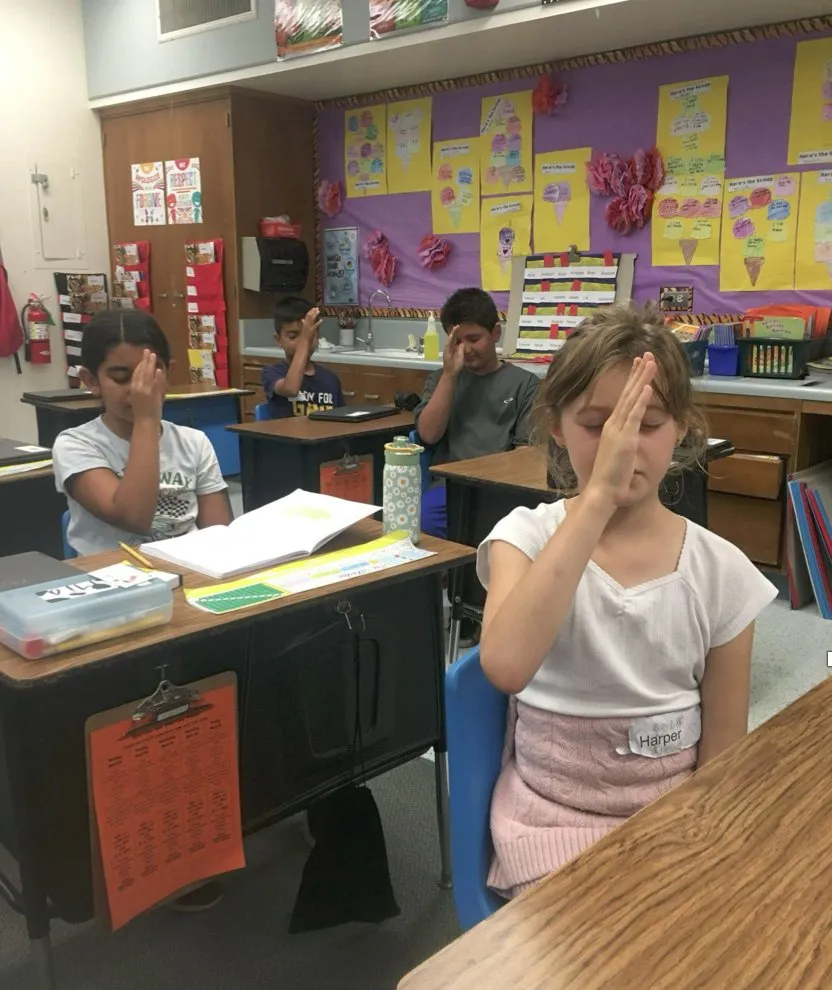 A class follows a guided practice at their desks, eyes closed and hands raised