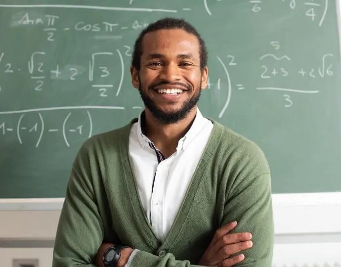 A smiling teacher stands with arms crossed in front of a chalkboard