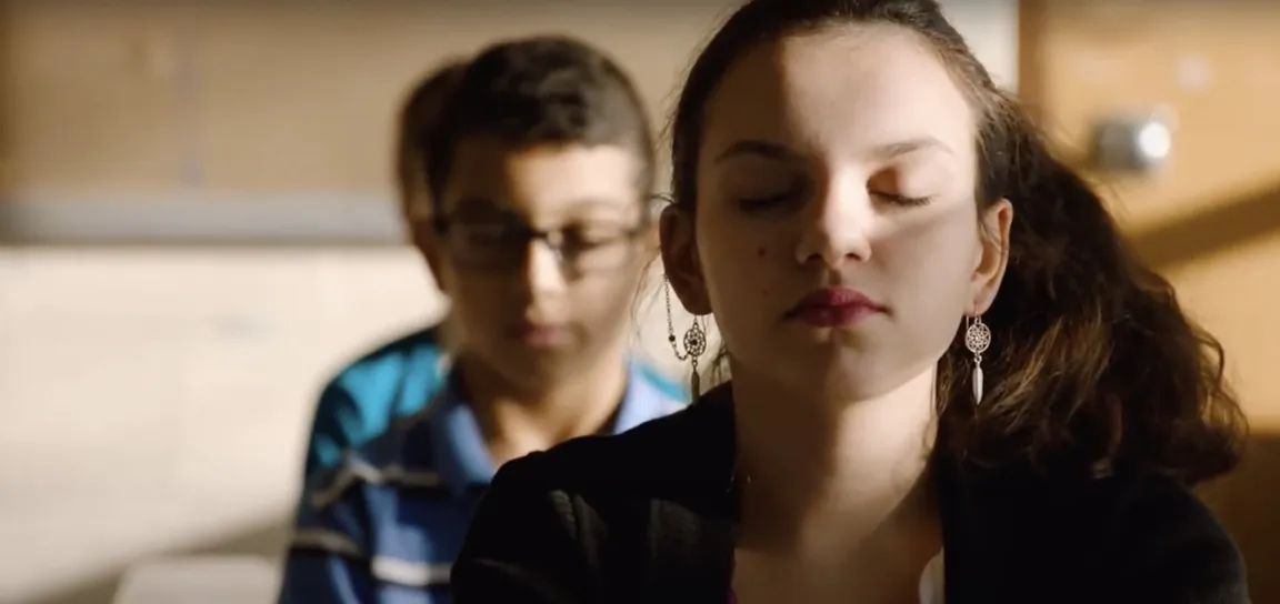 A student sits with eyes closed in a sunlit classroom during a daily mindfulness practice