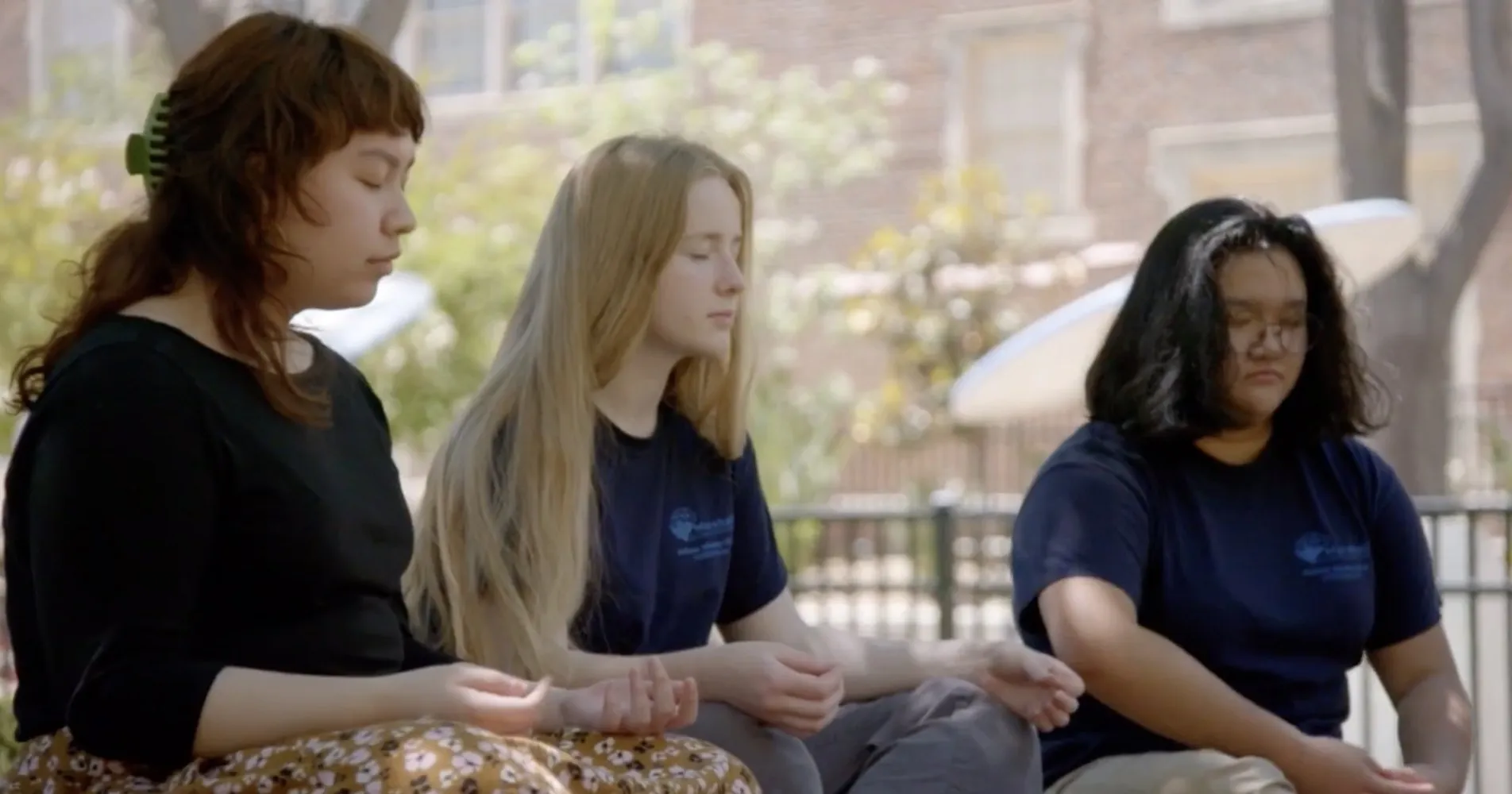 Three high school students sit outside with their eyes closed during a mindfulness practice