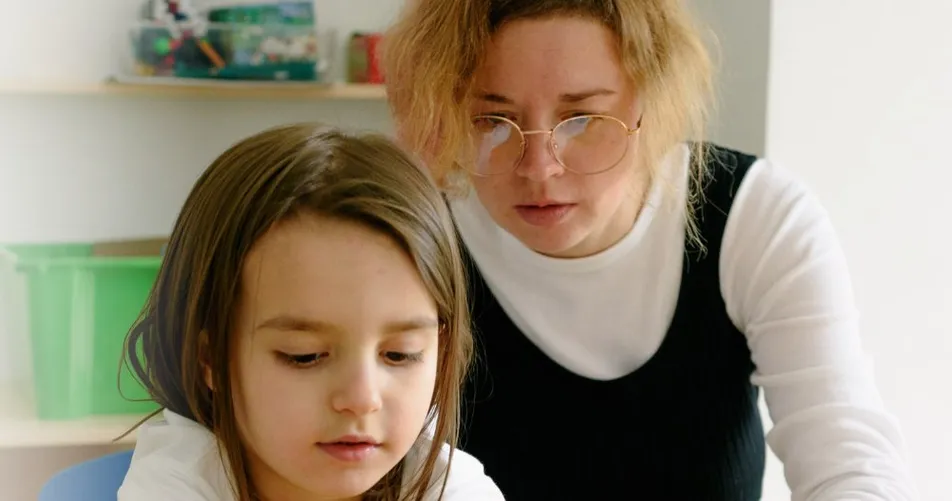 A teacher leans over a young girl’s shoulder as she works at her desk